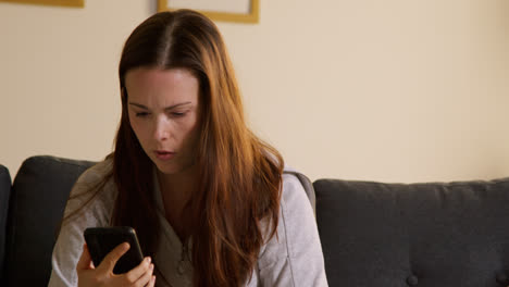 Anxious-Woman-Sitting-On-Sofa-At-Home-Looking-At-Mobile-Phone-Concerned-About-Social-Media-Or-Bad-News